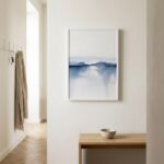 Abstract watercolour painting with soft blue tones depicting misty lake landscape in white frame on neutral wall